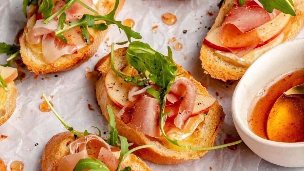 Slices of toasted bread topped with prosciutto, apple slices, arugula, and black pepper are arranged on parchment paper beside a small bowl of honey.