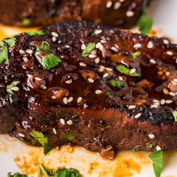 A cooked steak topped with glaze, sesame seeds, and chopped herbs sits on a white plate with sauce spread around.