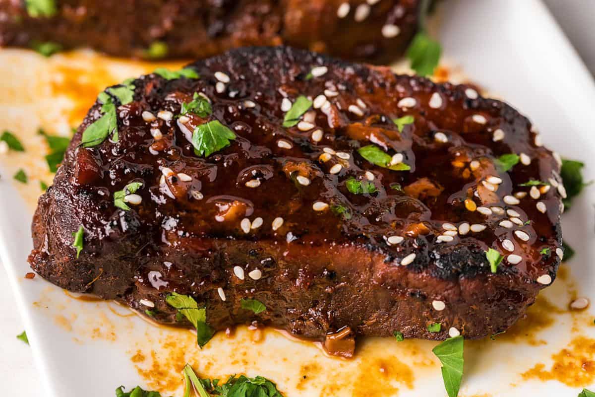A cooked steak topped with glaze, sesame seeds, and chopped herbs sits on a white plate with sauce spread around.