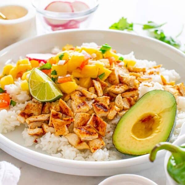 A plate of white rice topped with diced grilled chicken, mango salsa, half an avocado, and a lime wedge. In the background, fresh ingredients and garnishes are visible.