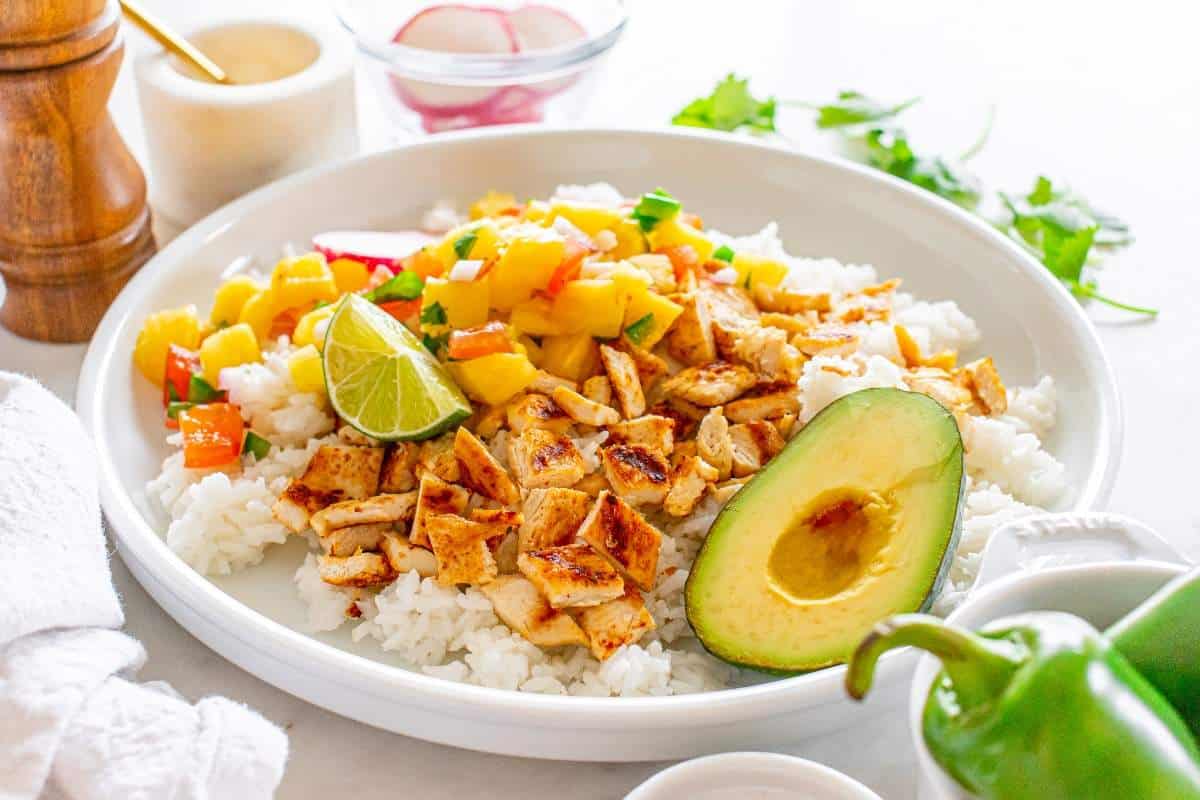 A plate of white rice topped with diced grilled chicken, mango salsa, half an avocado, and a lime wedge. In the background, fresh ingredients and garnishes are visible.