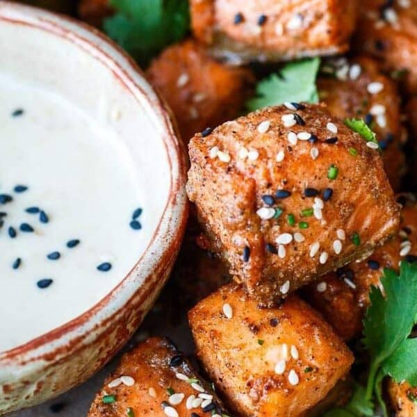 A bowl of white dipping sauce topped with black sesame seeds sits beside bite-sized pieces of seasoned salmon, which are garnished with additional sesame seeds and fresh cilantro leaves.