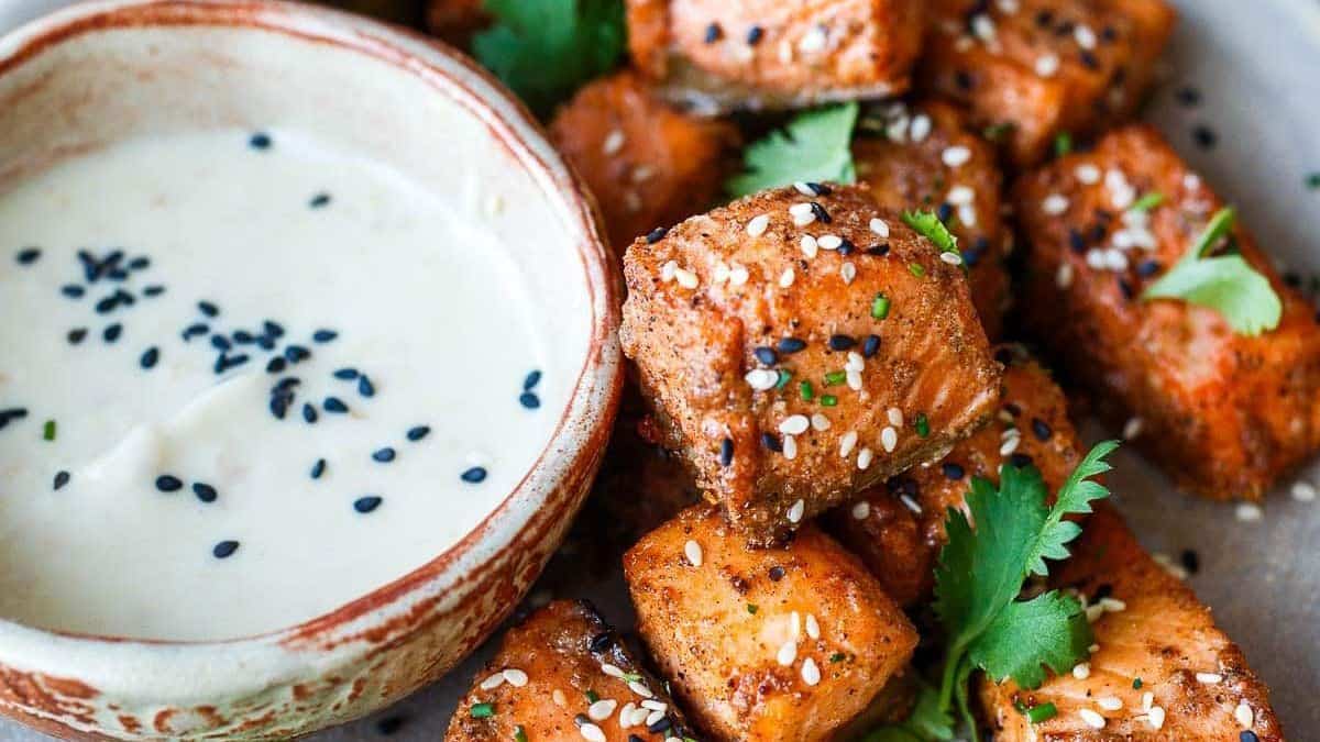 A bowl of white dipping sauce topped with black sesame seeds sits beside bite-sized pieces of seasoned salmon, which are garnished with additional sesame seeds and fresh cilantro leaves.