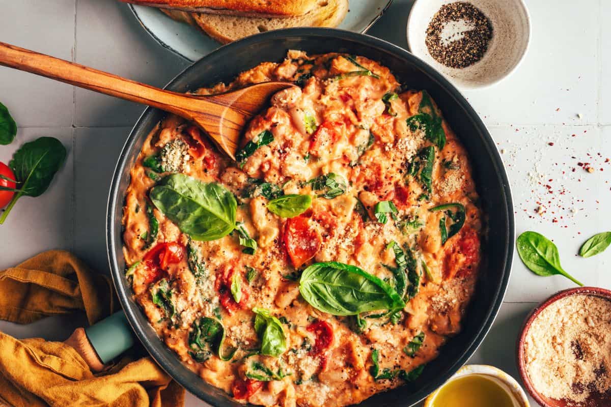 A skillet filled with creamy tomato spinach pasta topped with fresh basil leaves and grated cheese. A wooden spoon rests in the skillet. Surrounding the skillet are bread slices, a bowl of pepper, and various herbs and ingredients.