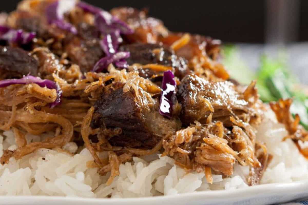 Close-up of a plate of white rice topped with shredded beef or pork, garnished with purple cabbage. The meat appears well-cooked with a slightly crispy texture. A side of greens is blurred in the background.