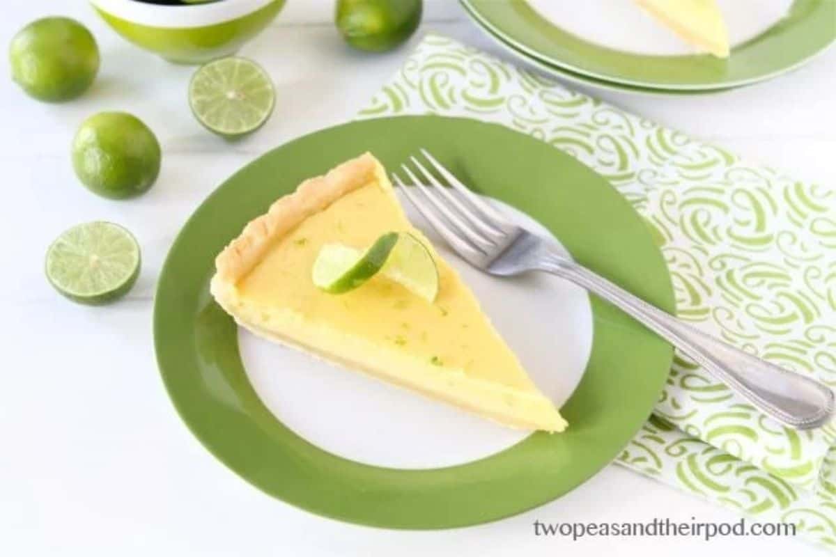 A slice of key lime pie garnished with a lime wedge sits on a white plate with a green rim. A fork is placed beside the pie. The plate is on a patterned green napkin, and whole and halved limes are scattered around. The website "twopeasandtheirpod.com" is visible.