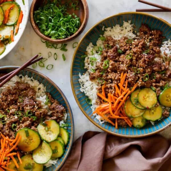 Two bowls of rice topped with ground meat, shredded carrots, and sliced cucumbers. A bowl of sliced cucumbers and carrots, a bowl of chopped green onions, chopsticks, and a fabric napkin are also visible on a white surface.