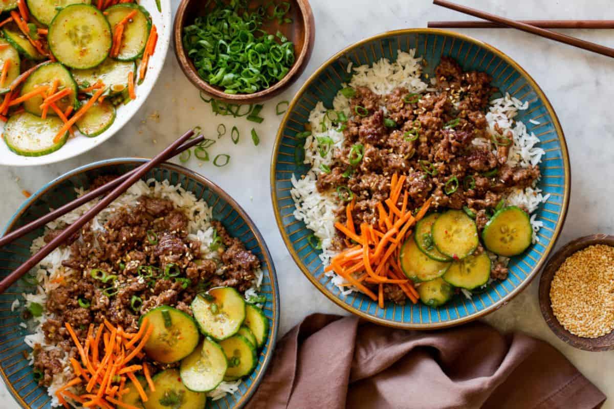 Two bowls of rice topped with ground meat, shredded carrots, and sliced cucumbers. A bowl of sliced cucumbers and carrots, a bowl of chopped green onions, chopsticks, and a fabric napkin are also visible on a white surface.