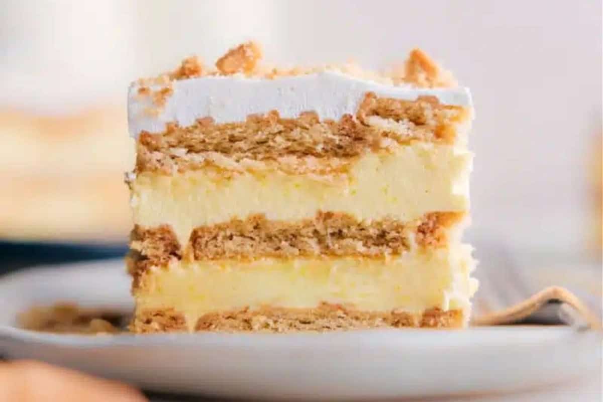 A close-up of a slice of layered cake on a white plate. The cake has layers of creamy yellow filling and crumbly golden crust, topped with a light layer of crushed crumbs. The background is softly blurred, focusing attention on the cake slice.