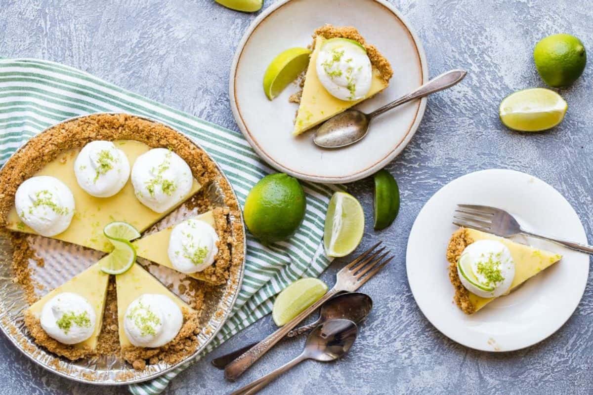 A key lime pie with six slices, garnished with whipped cream and lime zest. Two slices on plates with forks, limes, and spoons nearby, arranged on a striped towel and textured surface.