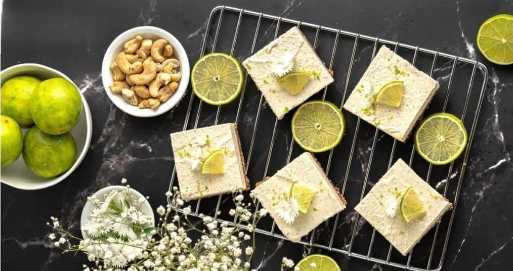 Image displaying "No Bake Vegan Key Lime Bars" on a cooling rack, garnished with lime slices and zest. A bowl of mixed nuts, fresh limes, and a small bouquet of white flowers are placed nearby on a black marble surface. 