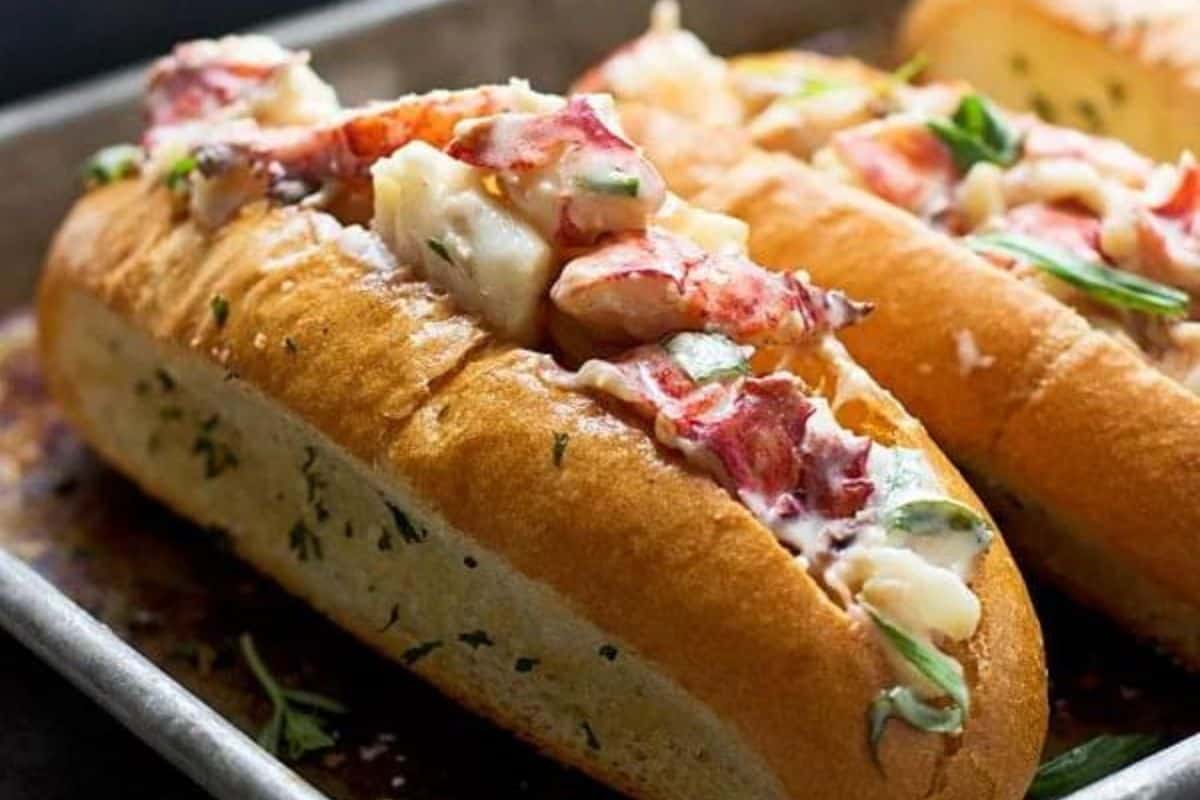 Two lobster rolls filled with chunks of lobster meat, garnished with herbs, are placed on a metal tray. The rolls are toasted to a golden brown, with visible pieces of lobster and creamy dressing spilling out slightly.