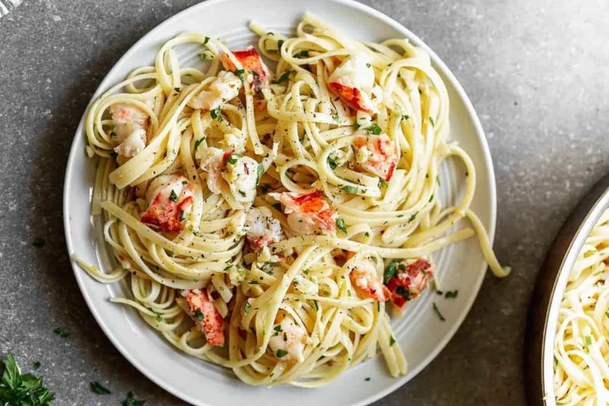 A plate of linguine pasta mixed with pieces of lobster meat, garnished with chopped parsley. The dish is presented on a white plate placed on a gray countertop, viewed from above. A partial view of a skillet with more pasta is visible on the side.
