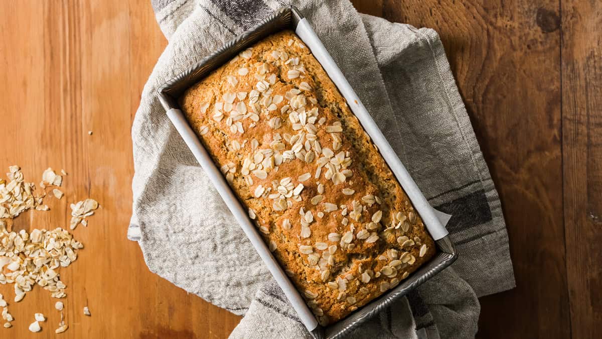A freshly baked loaf of oat bread rests in a rectangular metal baking tin, partially wrapped in a light-colored cloth. The bread is topped with scattered oats, and some oats have spilled onto the wooden table where the bread is placed.