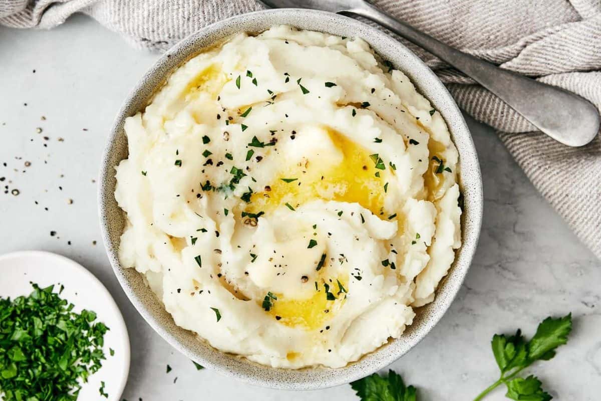 A bowl of creamy mashed potatoes garnished with melted butter, chopped parsley, and black pepper sits on a light gray surface. A small dish of chopped parsley and a sprig of fresh parsley are nearby. A gray striped cloth napkin and a spoon are in the background.
