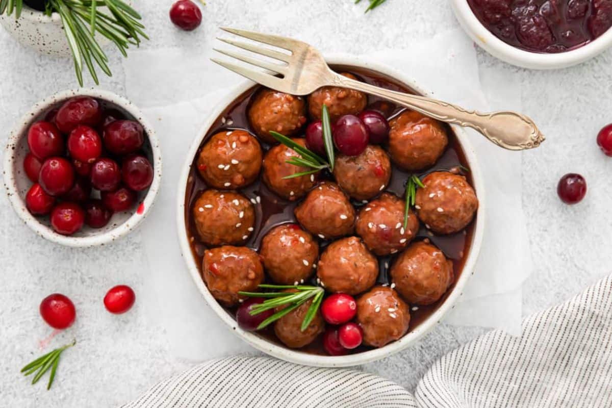 A plate of meatballs garnished with fresh rosemary and cranberries, with a fork resting on the plate. The dish is surrounded by additional bowls of cranberries and sauce on a light background, with a striped cloth at the bottom of the image.