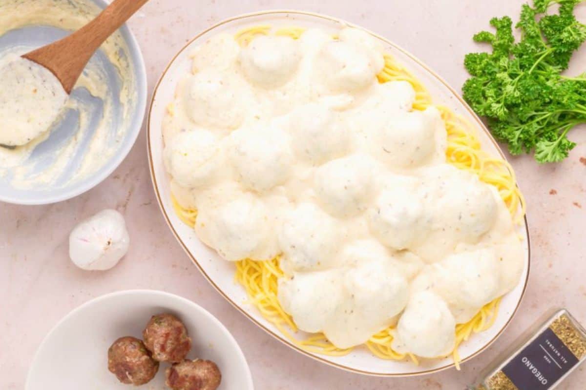 A dish of spaghetti topped with white cream sauce and meatballs on a white plate. A wooden spoon rests in a bowl of sauce on the side, with parsley and a jar of oregano nearby. Additional meatballs and a garlic clove are also placed around the dish.