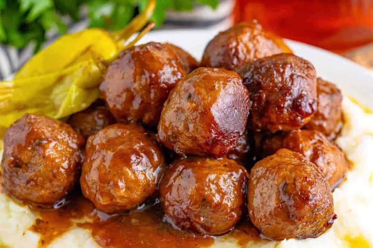A plate of glazed meatballs piled high on a bed of mashed potatoes, garnished with pickled peppers. The meatballs are coated in a shiny, dark brown sauce, and the mashed potatoes appear creamy and smooth. The background includes a hint of green foliage and a red drink.