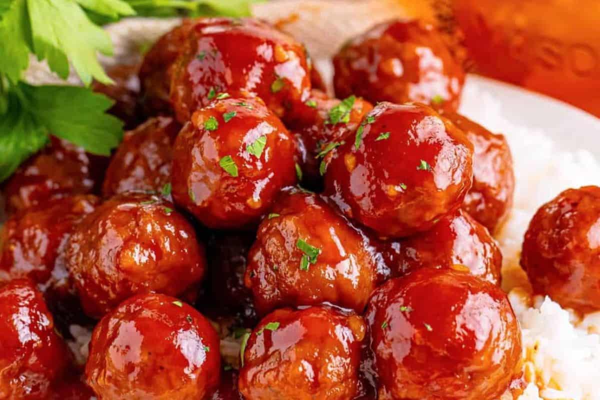A plate of glazed meatballs garnished with chopped parsley is shown. The meatballs are shiny and coated in a rich, brown sauce. Fresh parsley leaves are in the background, adding a touch of green to the vibrant and appetizing dish.