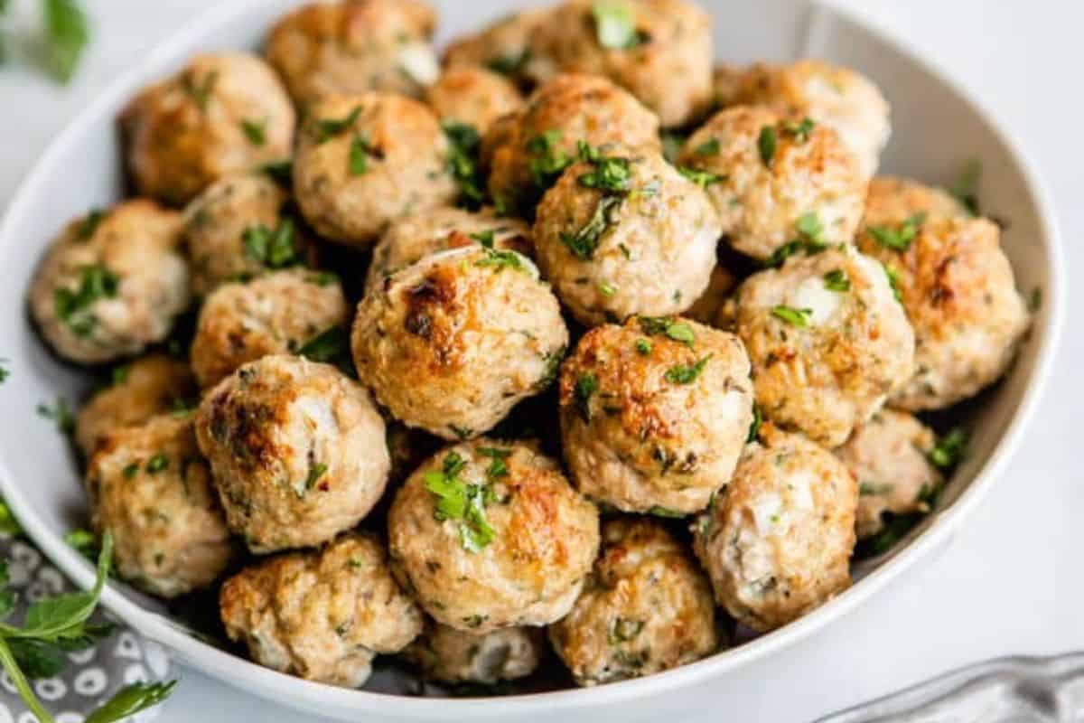 A white bowl filled with golden-brown meatballs garnished with chopped fresh parsley. The meatballs appear juicy and well-cooked, with a slightly crispy exterior. The close-up shot showcases the fresh herbs and appetizing texture of the meatballs.