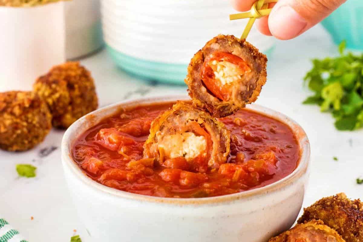 A hand holds a skewer of two breaded and fried mozzarella-stuffed meatballs, dipping them into a bowl of chunky tomato sauce. Fresh parsley and more meatballs are visible in the background. The scene is set on a white surface.