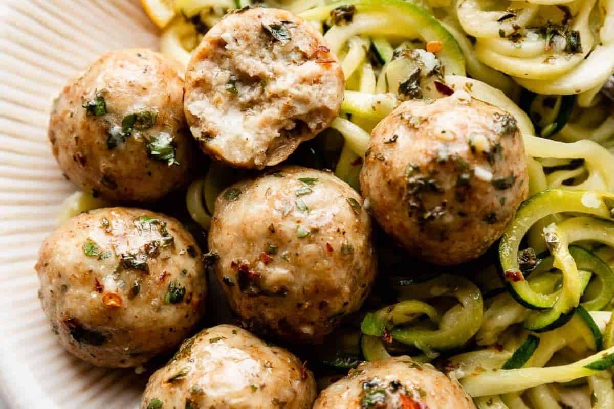 A plate of chicken meatballs, garnished with herbs and spices, served over a bed of spiralized zucchini noodles. One meatball is halved, displaying a juicy, well-cooked interior. The dish appears seasoned, vibrant, and delicious.