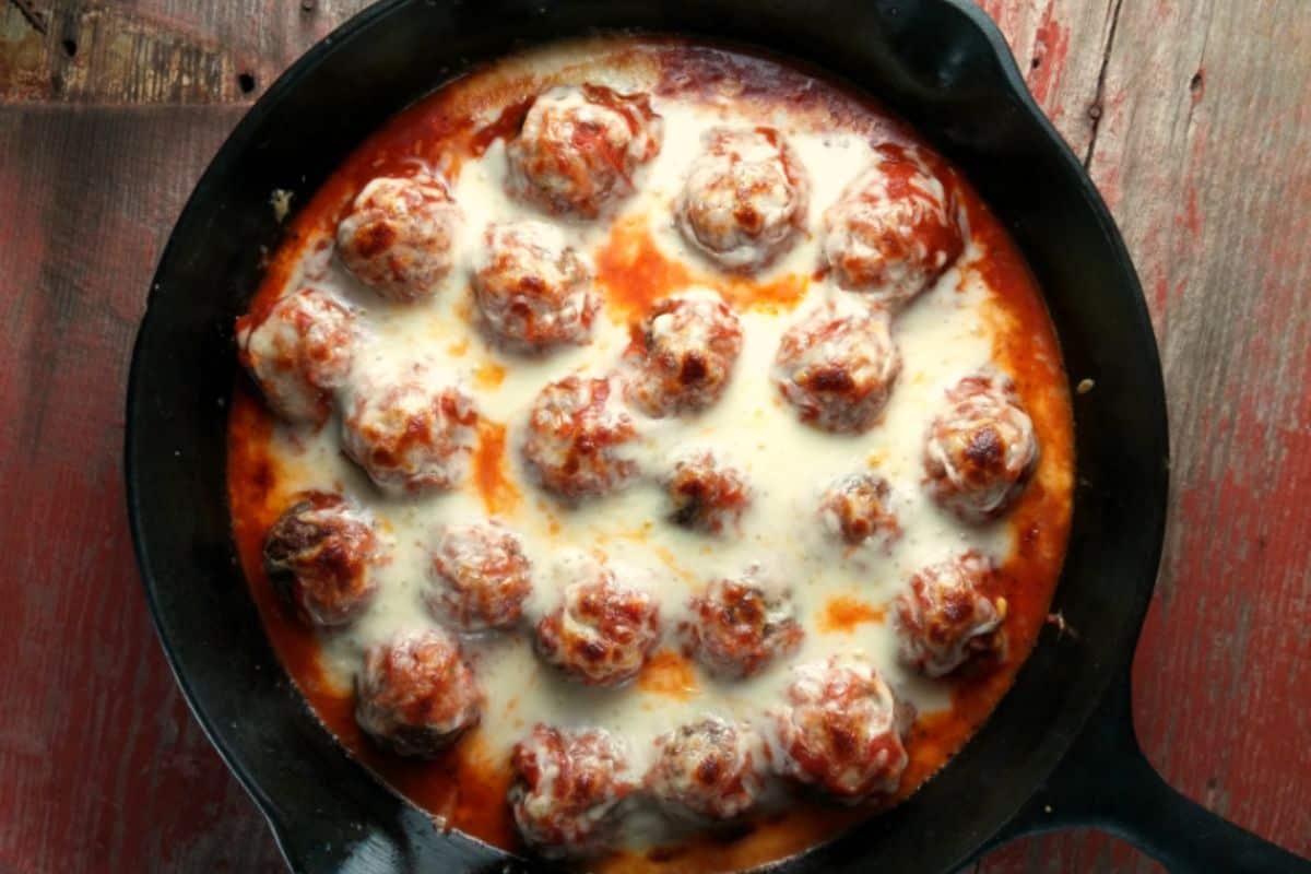 A cast-iron skillet filled with meatballs covered in melted cheese and marinara sauce. The skillet sits on a rustic, weathered wooden surface. The meatballs are evenly distributed and visibly baked with a crisp, cheesy topping.