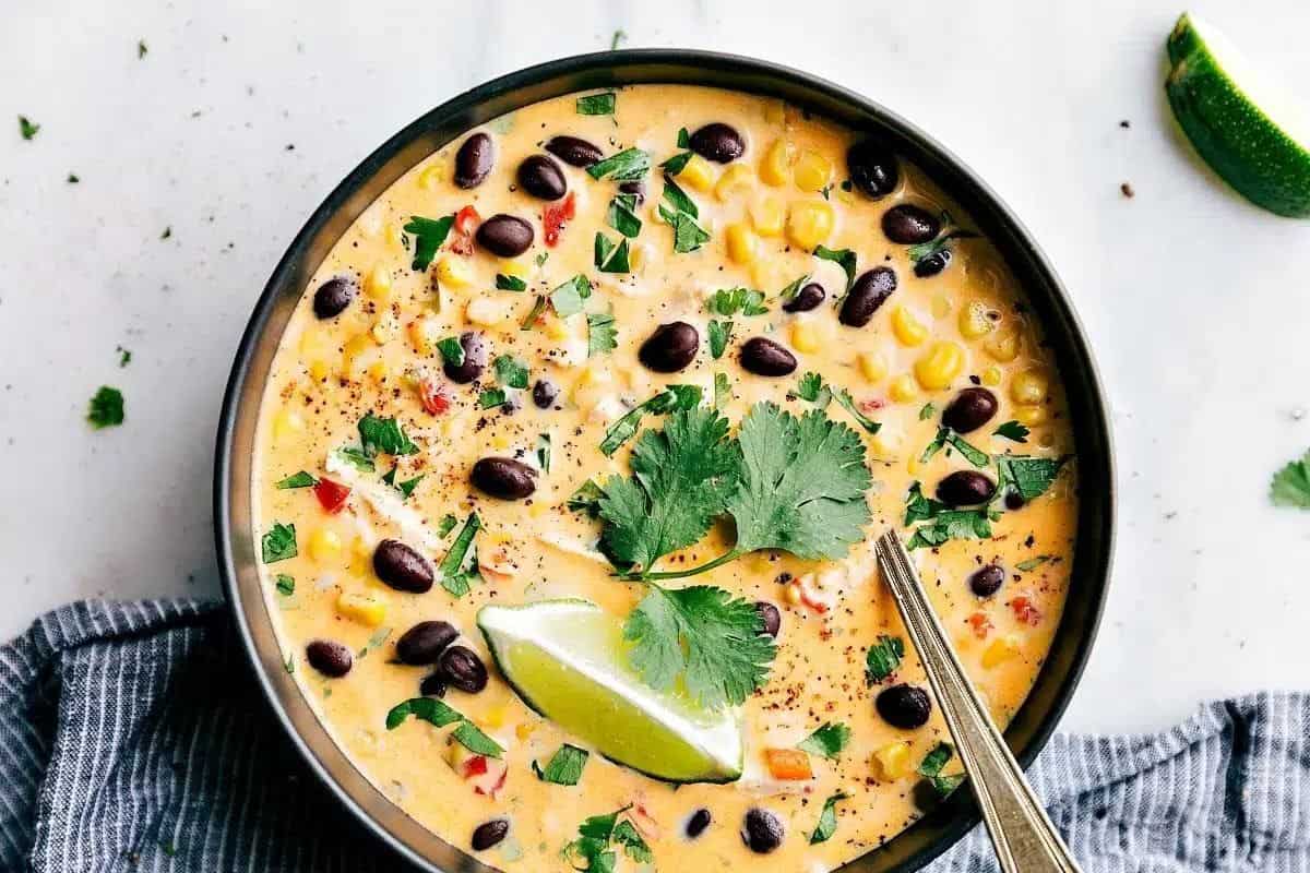 A bowl of creamy soup garnished with black beans, corn, cilantro, and a lime wedge. A metal spoon rests in the soup, and there is a piece of lime and cilantro garnish on the side. The bowl is placed on a gray cloth on a white surface.