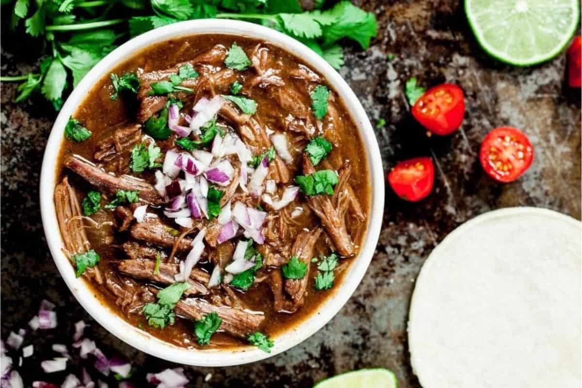 A bowl of beef birria stew garnished with chopped onions and cilantro, surrounded by lime wedges, chopped tomatoes, a tortilla, and fresh cilantro on a rustic surface.