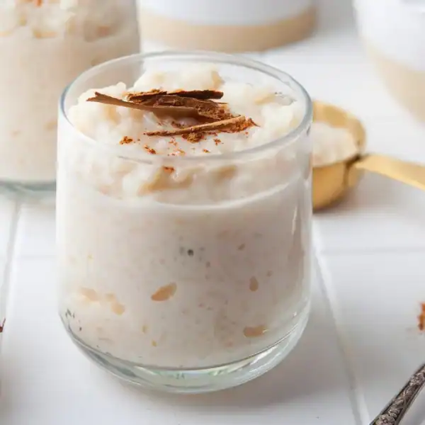 A glass of rice pudding garnished with a sprinkle of cinnamon and a cinnamon stick on top. A spoon with some cinnamon powder is lying next to it on a white table. Other cups and bowls are blurred in the background.