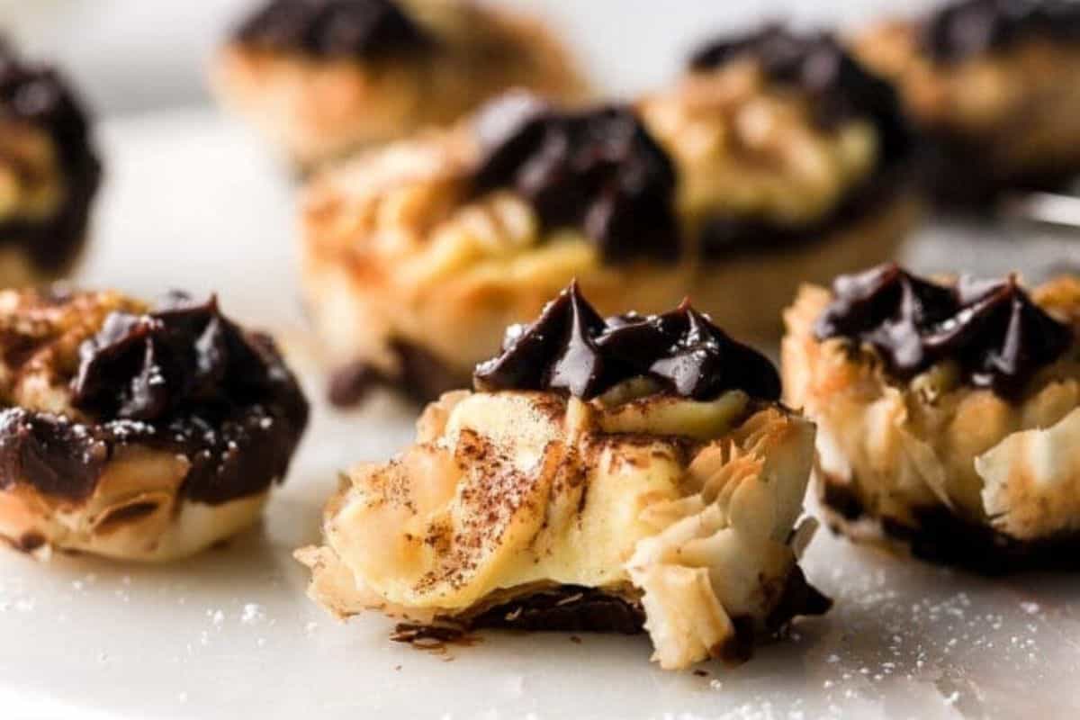 Close-up of bite-sized pastries topped with rich chocolate frosting. The pastries have a flaky, golden-brown crust, and some are dusted with a light sprinkle of powdered sugar. One pastry in the foreground has a visible bite taken out of it, revealing a creamy filling.