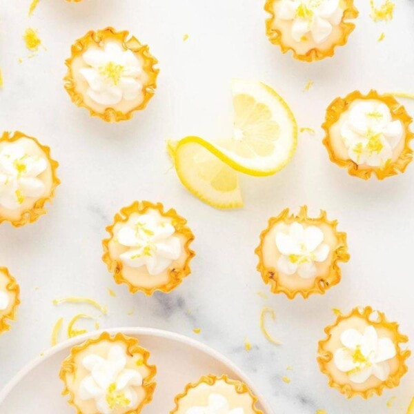 Mini lemon tarts with whipped cream and lemon zest garnish arranged on a white marble surface. The tarts have golden, crispy shells and decorative flowers made from the whipped cream. Slices of fresh lemon are scattered among the tarts for an added touch.