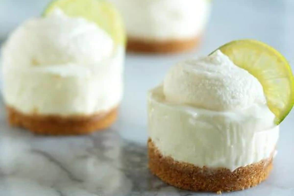 Miniature cheesecakes with a graham cracker crust, topped with whipped cream and garnished with a slice of lime on a marble surface.