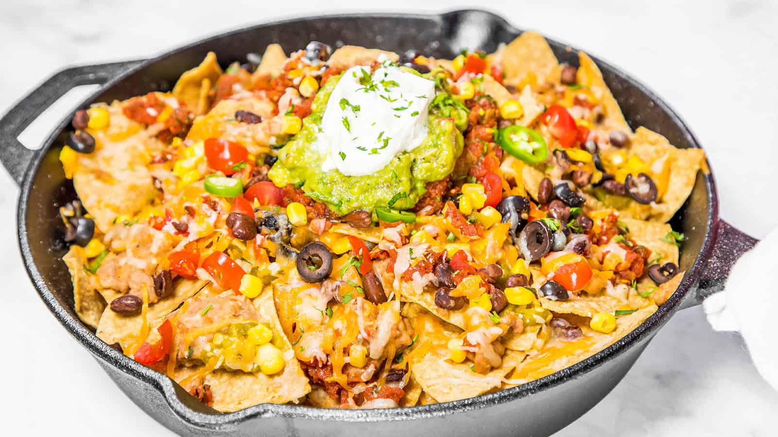 A skillet filled with loaded nachos featuring tortilla chips topped with melted cheese, diced tomatoes, olives, corn, black beans, jalapeño slices, and a dollop of guacamole and sour cream, garnished with chopped herbs.