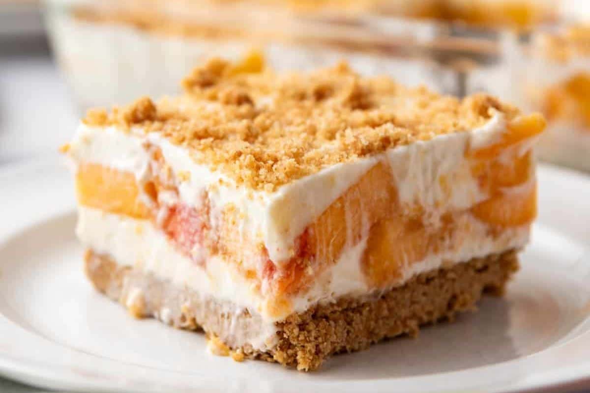 A slice of peach delight dessert sits on a white plate. The dessert features layers of creamy filling and peach slices, topped with a crumbly crust. Its topmost layer has a golden-brown crumble, adding texture and contrast to the creamy and fruity layers below.
