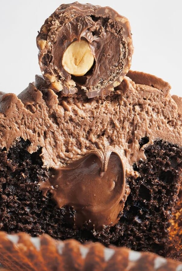 A chocolate cupcake cut in half, showing a Nutella filling inside, topped with creamy chocolate frosting and a chocolate-covered hazelnut candy.