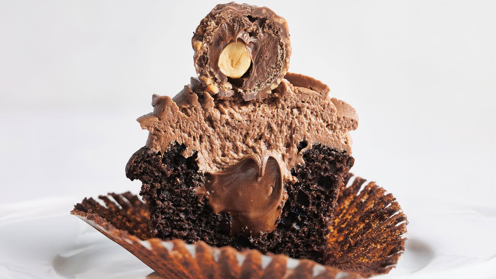 A close-up of a chocolate cupcake with its wrapper partially peeled. The cupcake is topped with creamy chocolate frosting, and half of a Ferrero Rocher chocolate is placed on top, revealing a hazelnut center. The white background contrasts with the rich, dark cupcake.