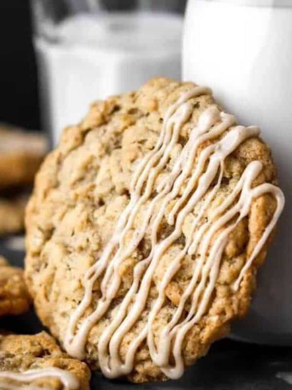 A large oatmeal cookie with white icing drizzle leans against a glass of milk, with more cookies and another glass in the background.