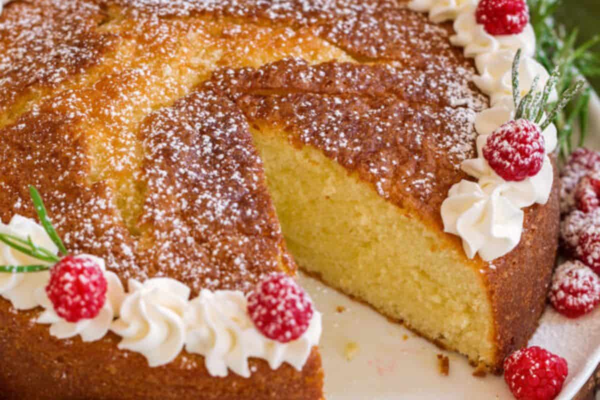 A golden-yellow cake with a slice cut out is decorated with swirls of white frosting, red raspberries, and sprigs of rosemary. The cake is dusted with powdered sugar and sits on a white plate.