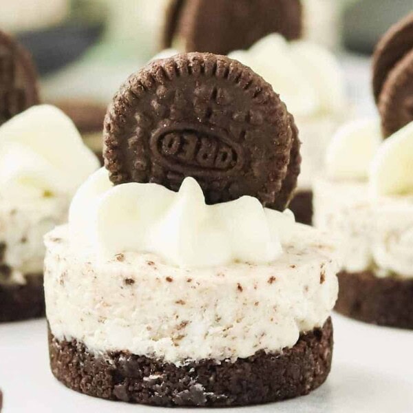 Mini cheesecakes topped with whipped cream and embellished with whole chocolate sandwich cookies. The cheesecakes have a brown crust, a creamy filling with cookie crumbs, and are attractively arranged.