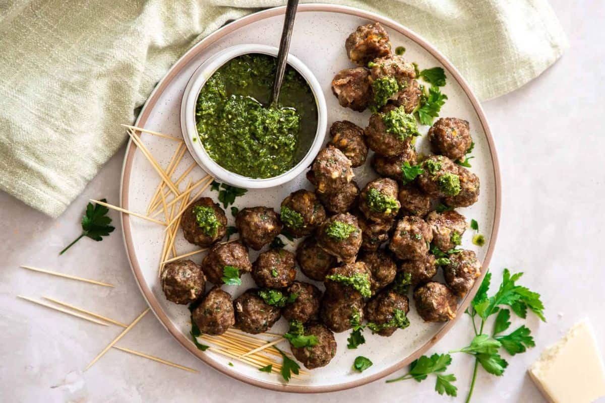 A round white plate filled with small meatballs garnished with green sauce, accompanied by a small bowl of green pesto and wooden toothpicks. Fresh parsley leaves are scattered around the plate, which is set on a light-colored surface with a green cloth nearby.