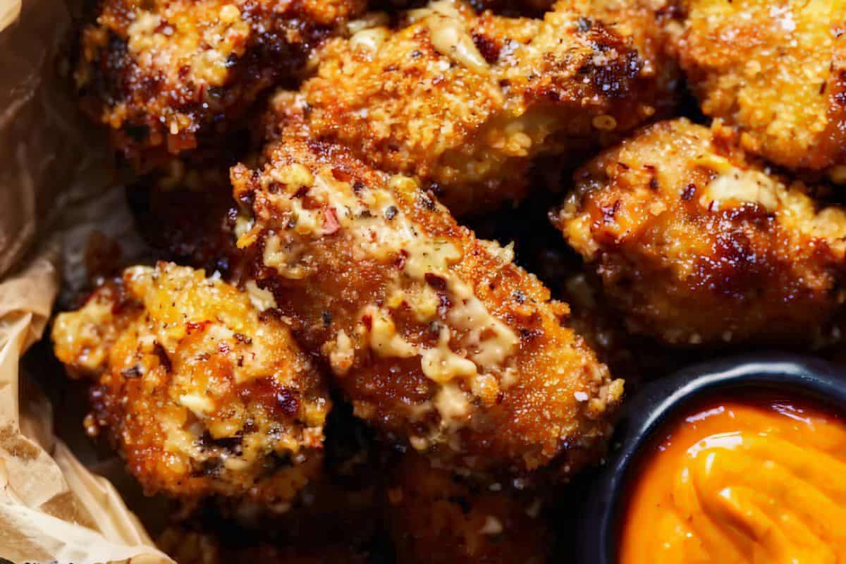 Close-up of crispy, golden-brown breaded chicken pieces drizzled with a creamy sauce, served in a rustic paper-lined basket. A small black bowl of vibrant orange dipping sauce is visible in the corner. The texture of the breading and sauce enhances the dish's appetizing look.