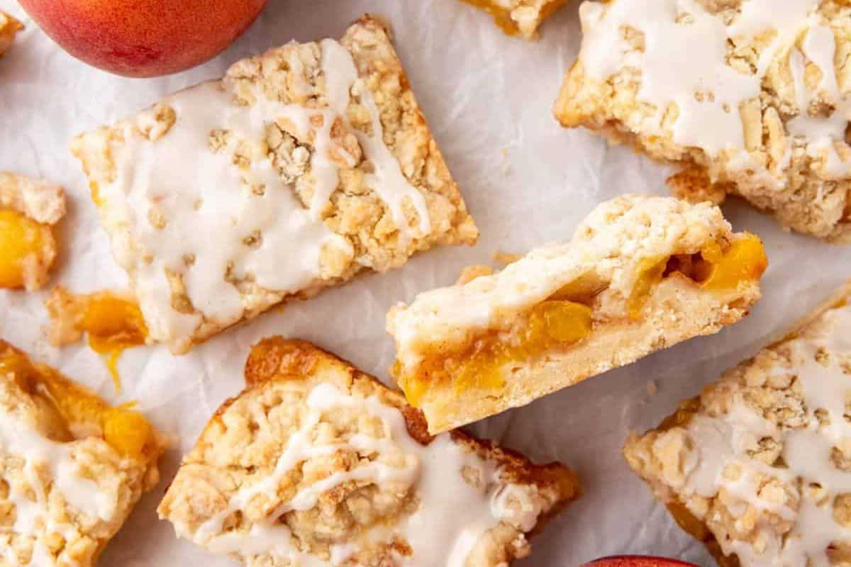 Close-up of several peach crumble bars, topped with a light glaze, scattered on parchment paper. The bars have a crumbly texture with visible peach filling layers. Two whole peaches are partially visible at the corners of the image.