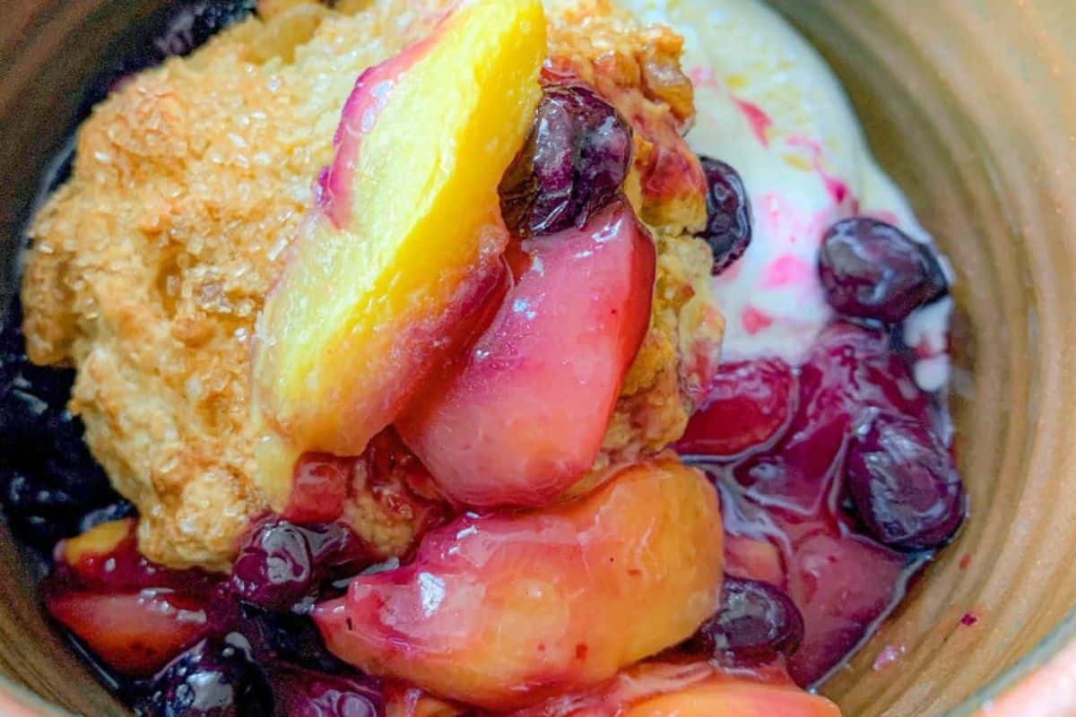 A close-up image of a fruity cobbler dessert. The dish features chunks of peaches, blueberries, and other mixed berries in a thick, syrupy sauce. There is a golden-brown biscuit topping, along with a dollop of vanilla ice cream that is starting to melt.