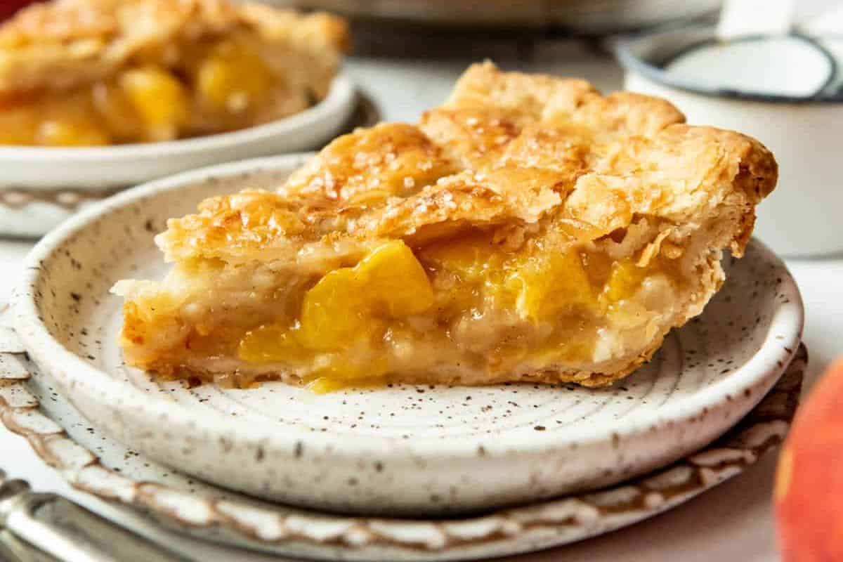 A close-up of a slice of peach pie on a speckled ceramic plate. The pie has a golden, flaky crust and is filled with chunks of juicy peaches. Two more slices of pie are visible in the blurred background.