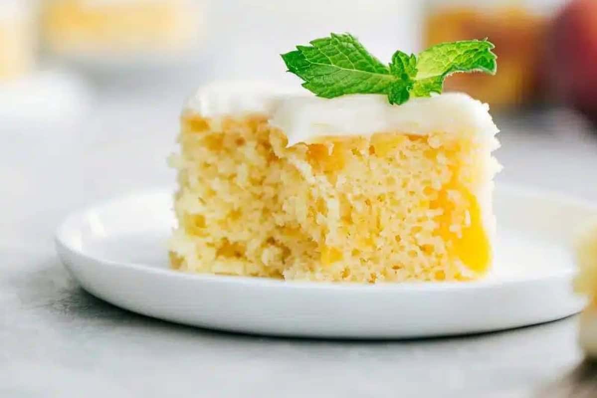 A slice of moist, yellow cake topped with white frosting and garnished with a sprig of mint on a white plate. The background is blurry, highlighting the cake's texture and garnishing.