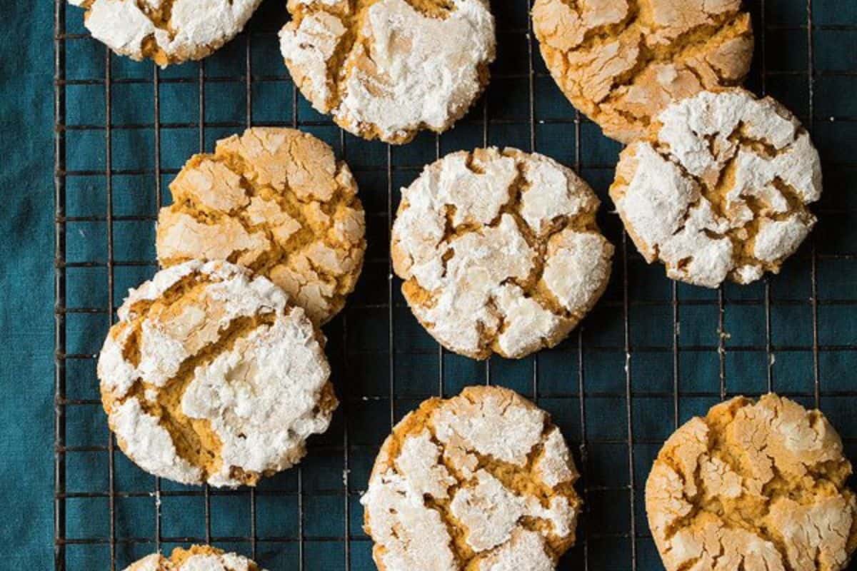 A cooling rack with several round, cracked cookies. Some cookies are dusted with powdered sugar, while others show a golden-brown, crinkled surface. The background is a dark, textured fabric.