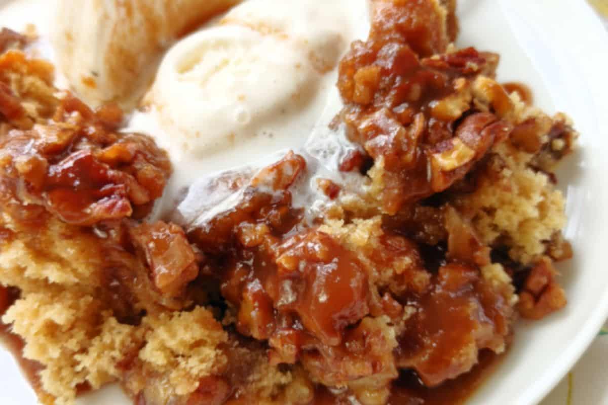 A close-up image of a dessert featuring a serving of caramel pecan bread pudding. The pudding has a light, crumbly texture and is topped with a rich, caramel sauce and bits of pecan. In the background, there is a scoop of vanilla ice cream starting to melt.