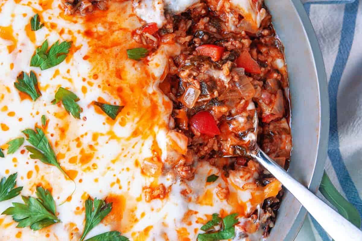 Close-up of a dish of moussaka, showcasing a creamy béchamel sauce topped with fresh parsley leaves on the left half. The right half is partially scooped out, revealing an interior of minced meat, tomatoes, and spices. A spoon is resting in the scooped-out section.