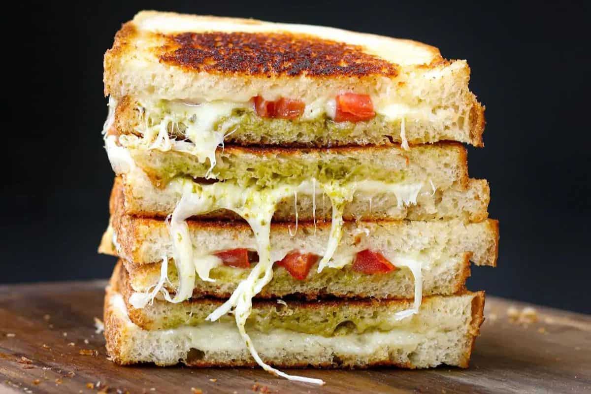 A stack of four grilled cheese sandwiches layered with melted cheese, tomato slices, and a green pesto spread on toasted bread. The sandwiches are placed on a wooden surface against a dark background.